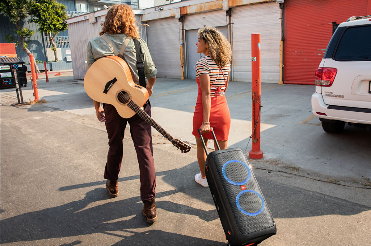 Woman wheeling a JBL Partybox 310 down a street next to a guy with a guitar